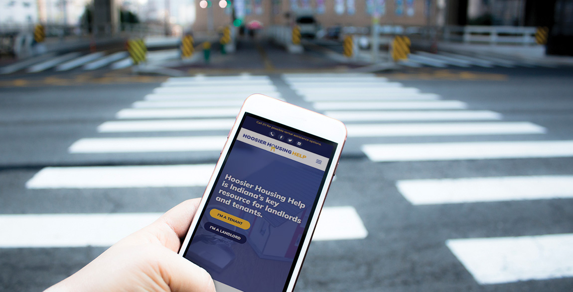 A person's hand holding a phone showing the Hoosier Housing Help website in the foreground with a crosswalk on a street in the background.