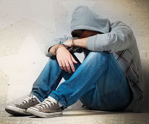 A handcuffed teen sits on with his arms covering his face.