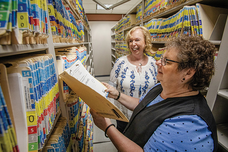 Clerks smile while standing in amid shelves of old paper records