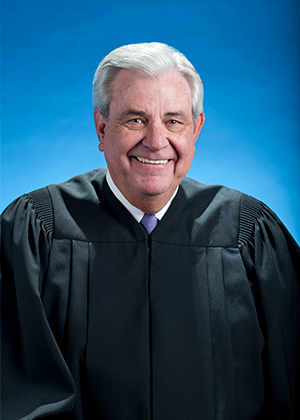 Color head shot of Judge Michel Barnes wearing black judges' robes, in front of blue background.