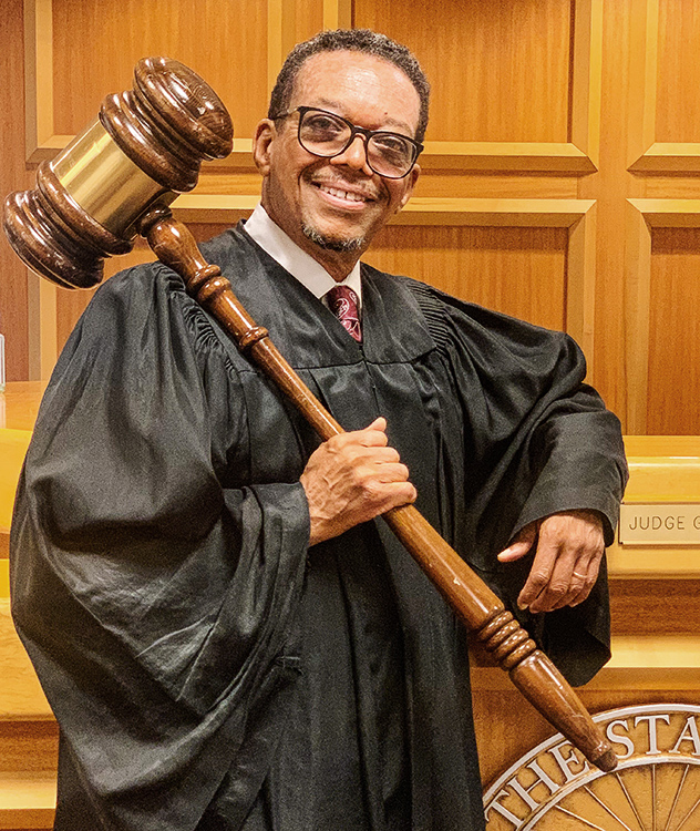 Judge Geoffrey Gaither holds an oversized gavel in front of a courtroom bench.