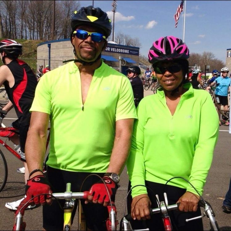 Judge Gaither and his wife wear fluorescent yellow shirts and helmets while straddling bicycles.