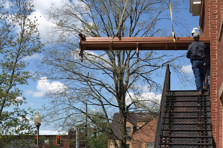 A pew extends out of a doorway on the second floor of a building. A worker guides the pew out. Crane straps support the weight of the pew.