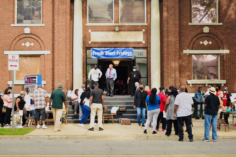 People crowd a sidewalk outside of a brick church. A blue banner hangs over the steps of the entryway reads, "Fresh Start Friday."