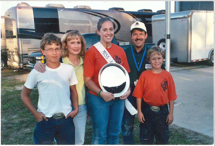 Daytime. Judge Christofeno and his family pose for a photo in front of an RV. His daughter holds a shine round award and wears a sash. Everyone wears jeans and short-sleeve shirts.