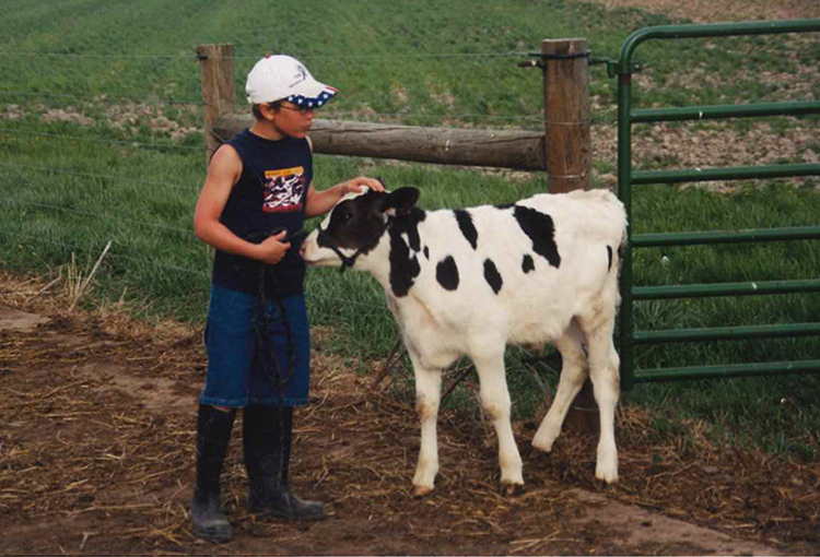 A boy in a baseball hat holds a lead attached to a calf. He pets the head of the calf and it presses its nose against the boy.