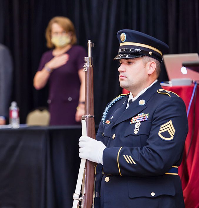 Armed service member stands at attention; Chief Justice, in the background, saying the pledge of allegiance.