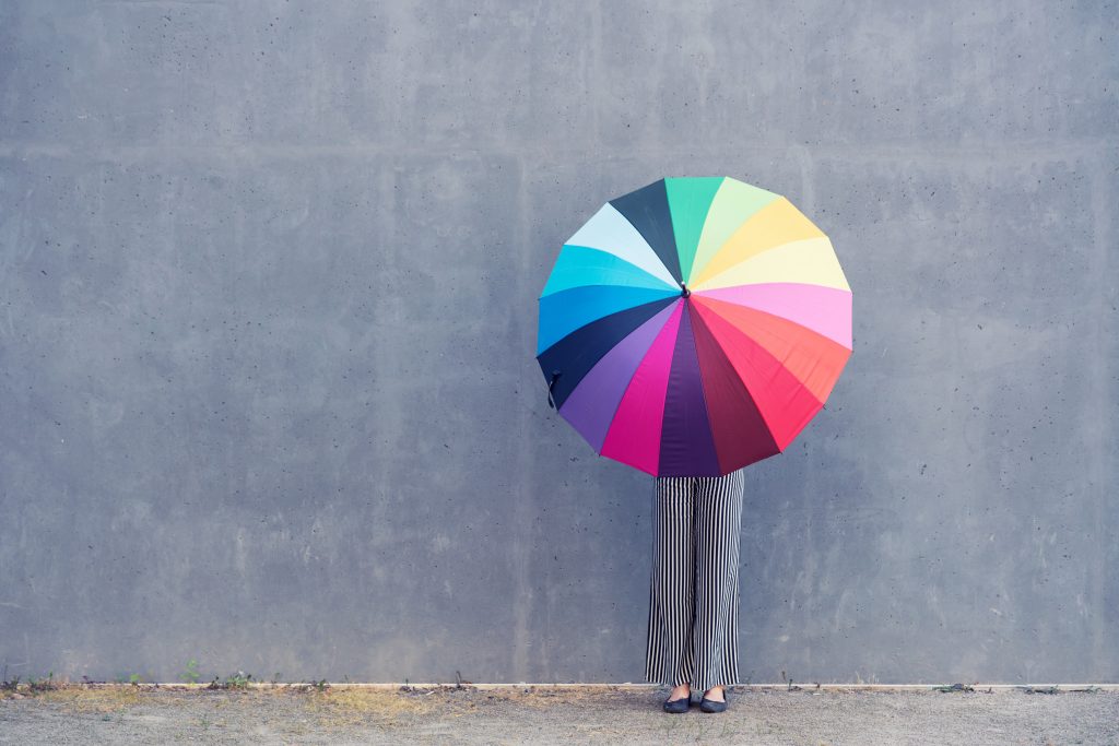 Anonymous person standing against a wall holding a colorful umbrella in front of her, blocking our view of her.