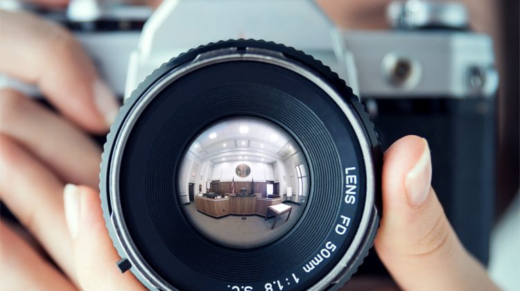 Woman holding camera pointed at the viewer with a courtroom reflected in the camera lens.