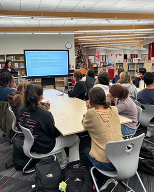 Students gather around tables in a school library and listen as Magistrate Garza addresses them.