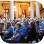 Volunteers in blue shirts sit and listen to guest speakers at the Statehouse.