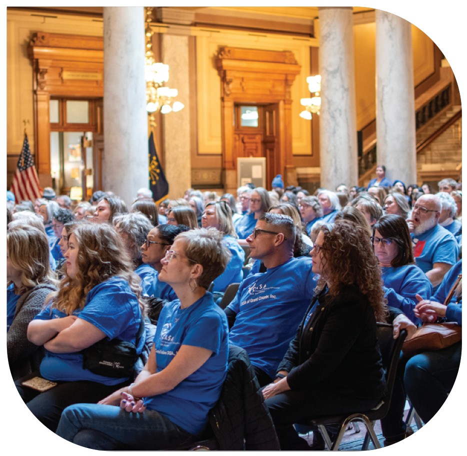 Volunteers in blue shirts sit and listen to guest speakers at the Statehouse.